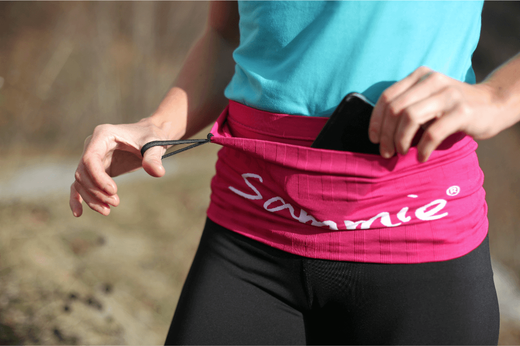 La running belt dans sa version rose portée par une sportive qui récupère à l'intérieur de la ceinture son téléphone portable